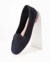 A single navy blue suede penny loafer shoe displayed at an angle on a white geometric block against a clean white background.